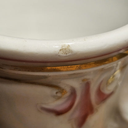 Gros plan sur le bord supérieur de la tasse à brûlot en faïence, montrant un petit éclat d'émail ainsi que le filet doré et le décor en relief rose, témoignant de l'ancienneté de la pièce.