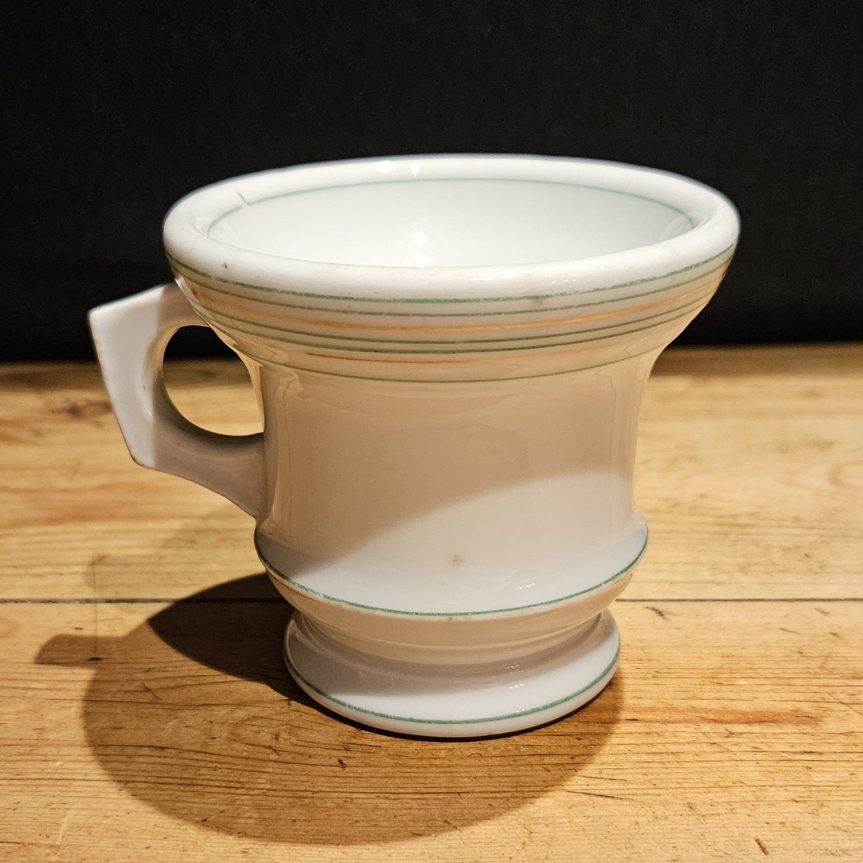 Vue de face de la tasse à brûlot en faïence blanche avec filets verts et or, anse rectangulaire sur le côté gauche, pied annulaire, posée sur une table en bois brut, fond noir.