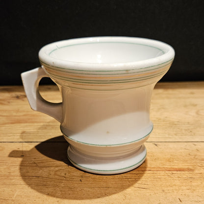 Vue de face de la tasse à brûlot en faïence blanche avec filets verts et or, anse rectangulaire sur le côté gauche, pied annulaire, posée sur une table en bois brut, fond noir.