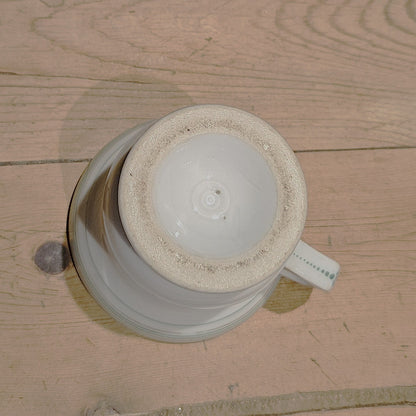 Vue du dessous de la tasse à brûlot en faïence blanche, montrant la base circulaire non émaillée avec une légère patine d'ancienneté, et l'anse rectangulaire avec motifs verts visible sur le côté.