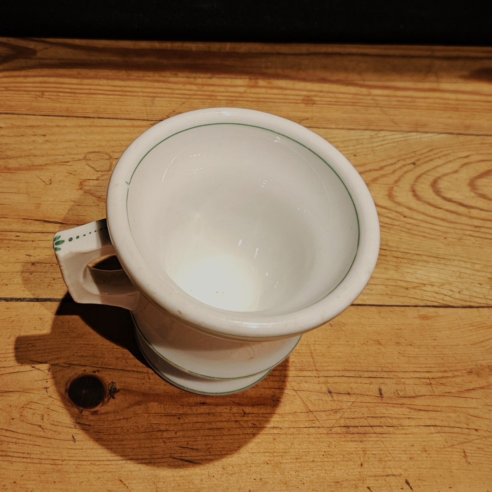 Vue plongeante de l'intérieur de la tasse à brûlot en faïence blanche, montrant l'émail blanc lisse, le bord avec filet vert, et l'anse rectangulaire, posée sur une table en bois brut.