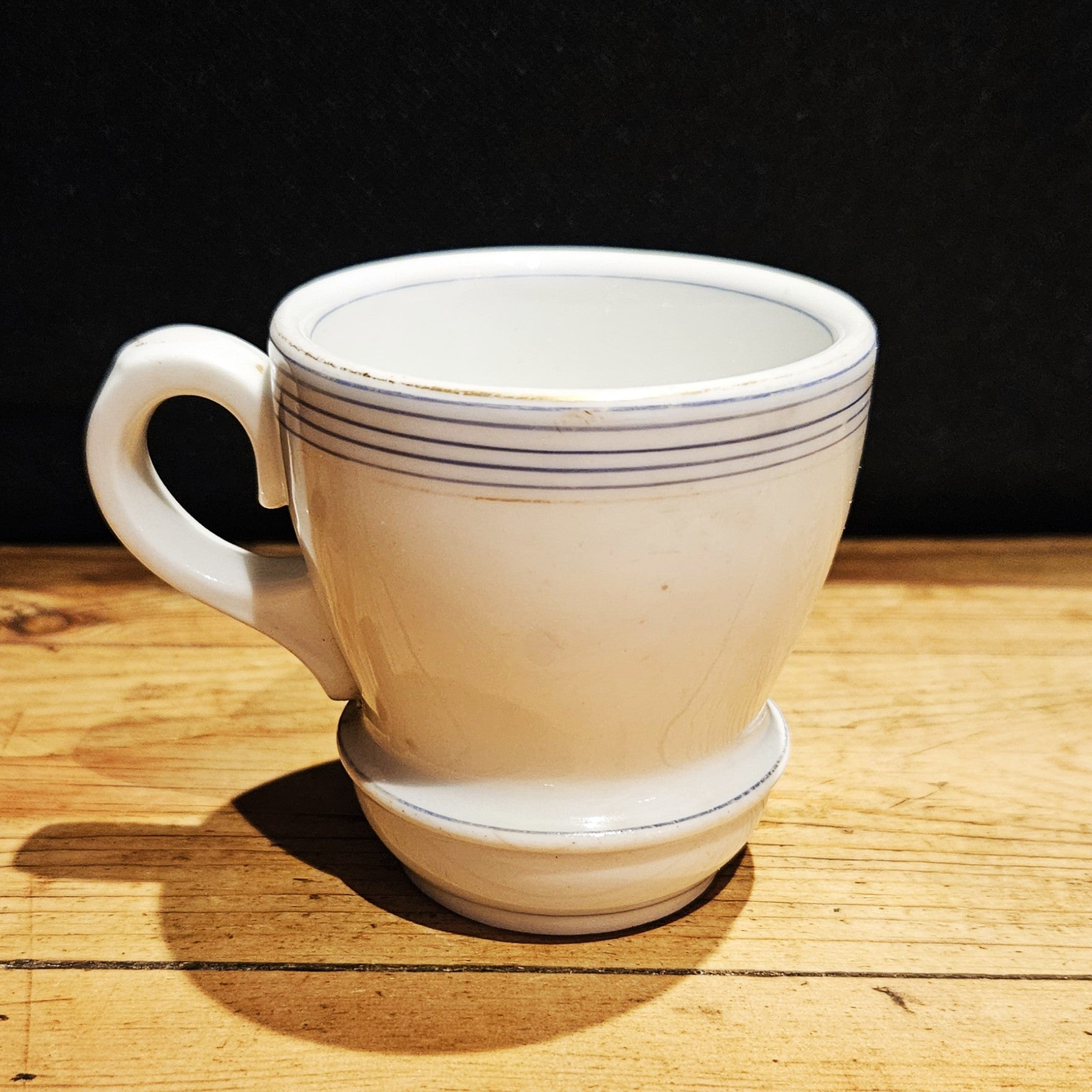 Tasse à brûlot ancienne en faïence blanche, forme tronconique sur pied , ornée de fins filets bleus sur le bord supérieur, anse ronde à gauche, posée sur une table en bois brut, fond noir.