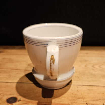 Vue arrière de la tasse à brûlot en faïence blanche, montrant l'anse ronde avec une légère usure dorée visible, et les fins filets bleus sur le bord supérieur, fond noir.