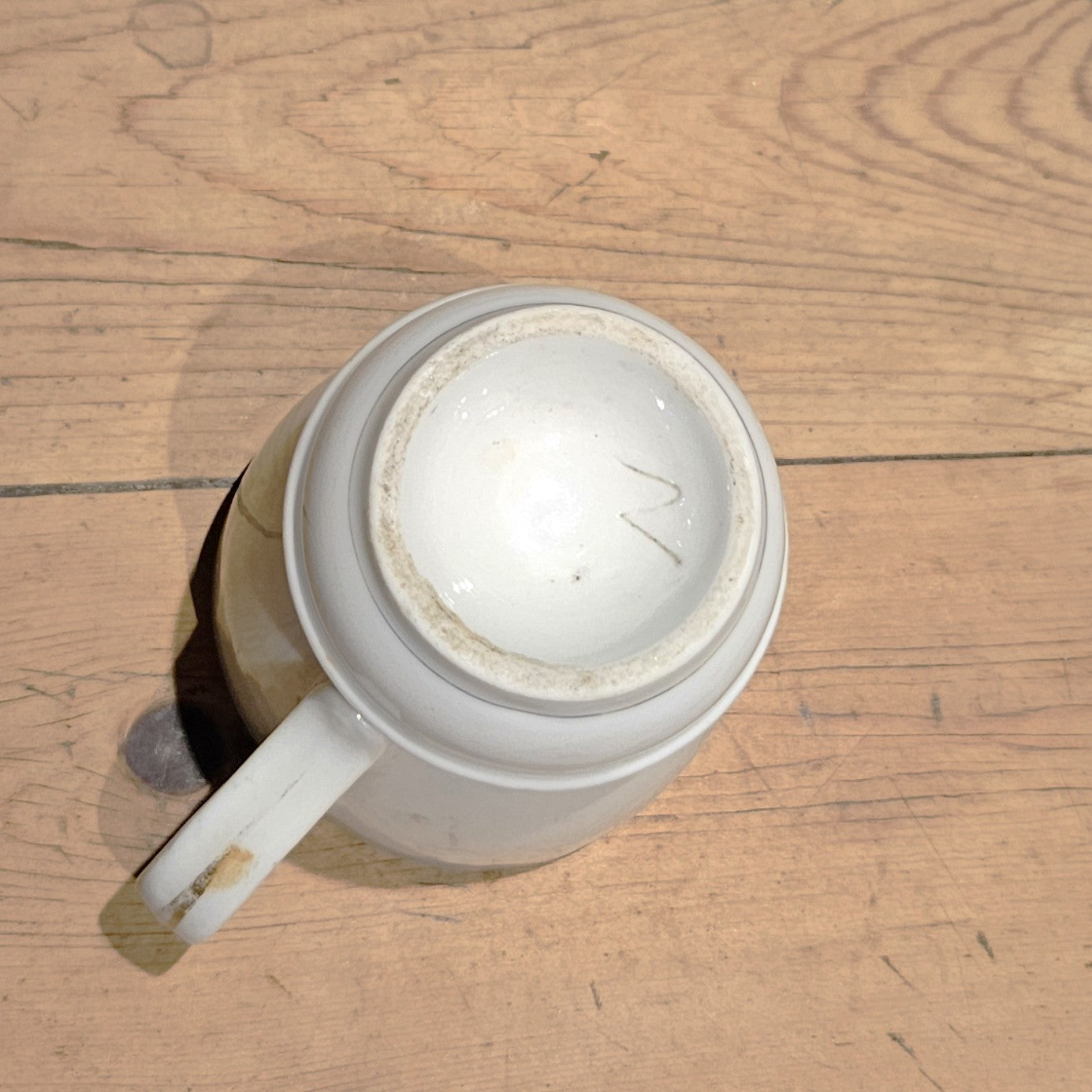 Vue du dessous de la tasse à brûlot en faïence blanche, montrant la base circulaire émaillée avec une marque de potier gravée en forme de lettre, et le pied bombé avec filet bleu