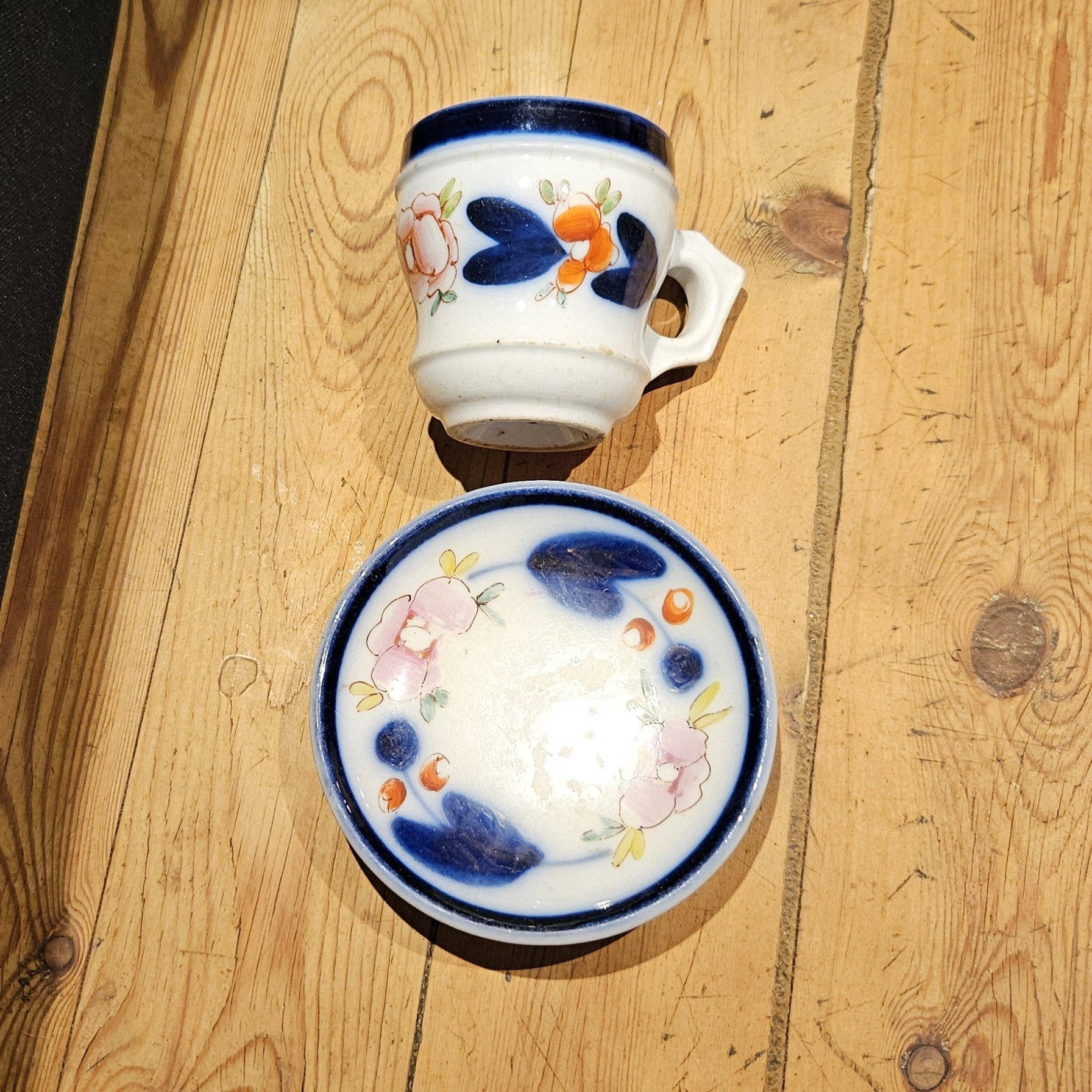 Vue plongeante d'une tasse à brûlot et sa soucoupe en faïence blanche, avec bandeau bleu cobalt et décor floral peint à la main (roses orangées et feuilles bleu-vert), posées sur un parquet en bois brut.