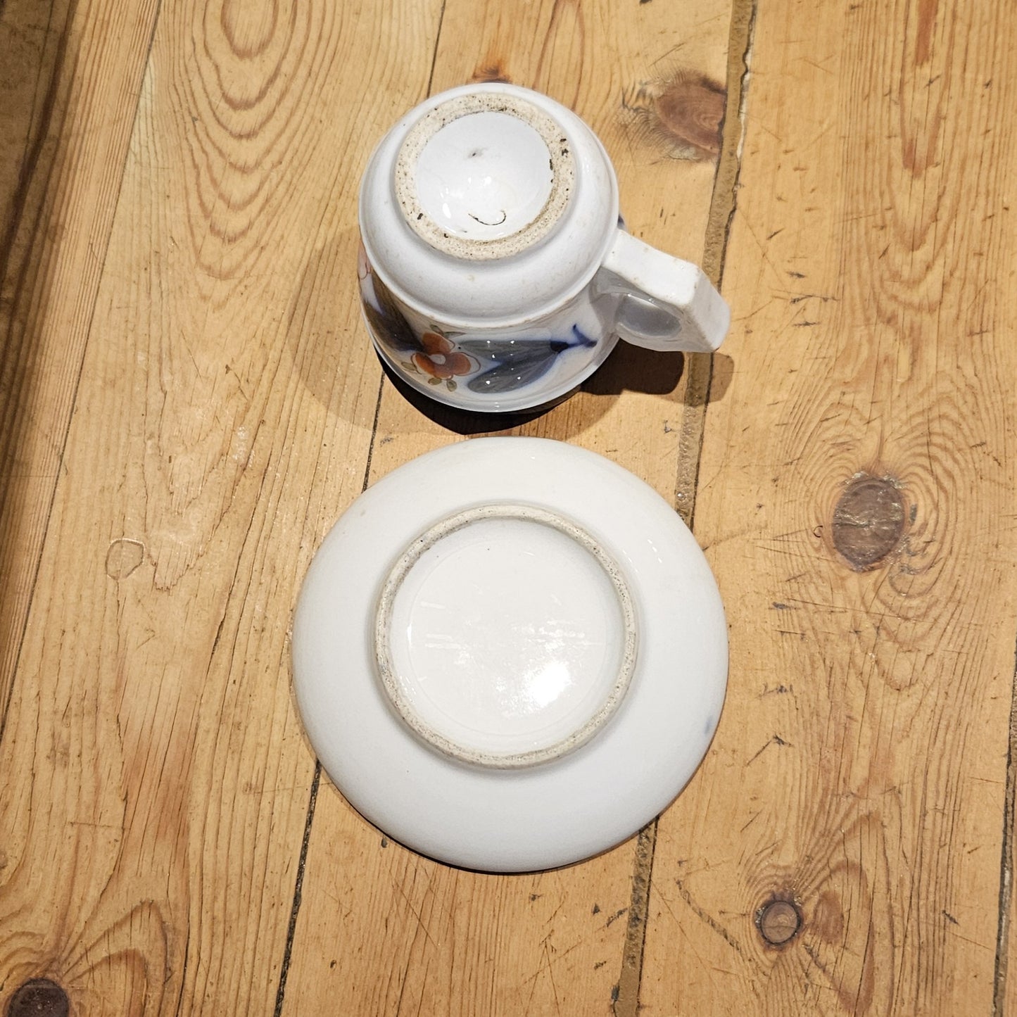 Vue du dessous de la tasse à brûlot et de la soucoupe en faïence blanche, montrant les bases partiellement émaillées avec patine d'ancienneté, sans marque de potier visible, posées sur un parquet en bois brut.