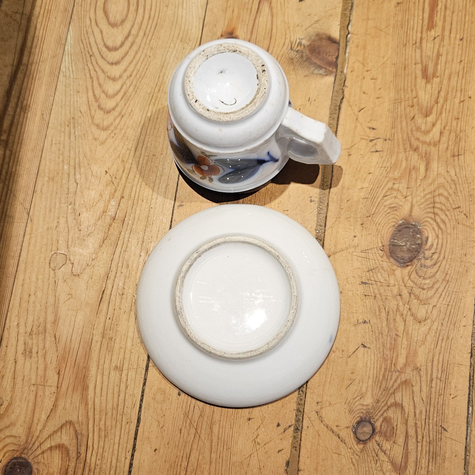 Vue du dessous de la tasse à brûlot et de la soucoupe en faïence blanche, montrant les bases partiellement émaillées avec patine d'ancienneté, sans marque de potier visible, posées sur un parquet en bois brut.