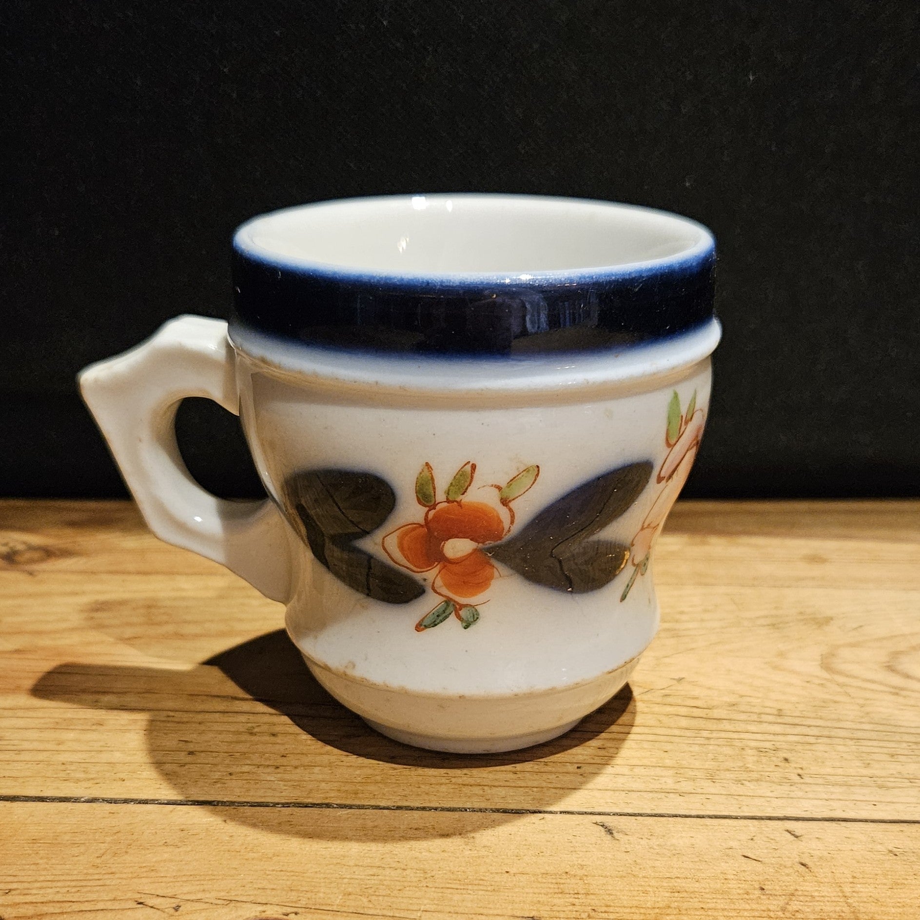 Tasse à brûlot ancienne en faïence blanche, avec large bandeau bleu cobalt sur le bord supérieur, décor floral peint à la main (fleurs orangées et feuilles sombres), anse  à gauche, posée sur une table en bois, fond noir.