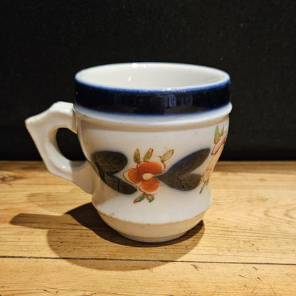 Tasse à brûlot ancienne en faïence blanche, avec large bandeau bleu cobalt sur le bord supérieur, décor floral peint à la main (fleurs orangées et feuilles sombres), anse  à gauche, posée sur une table en bois, fond noir.