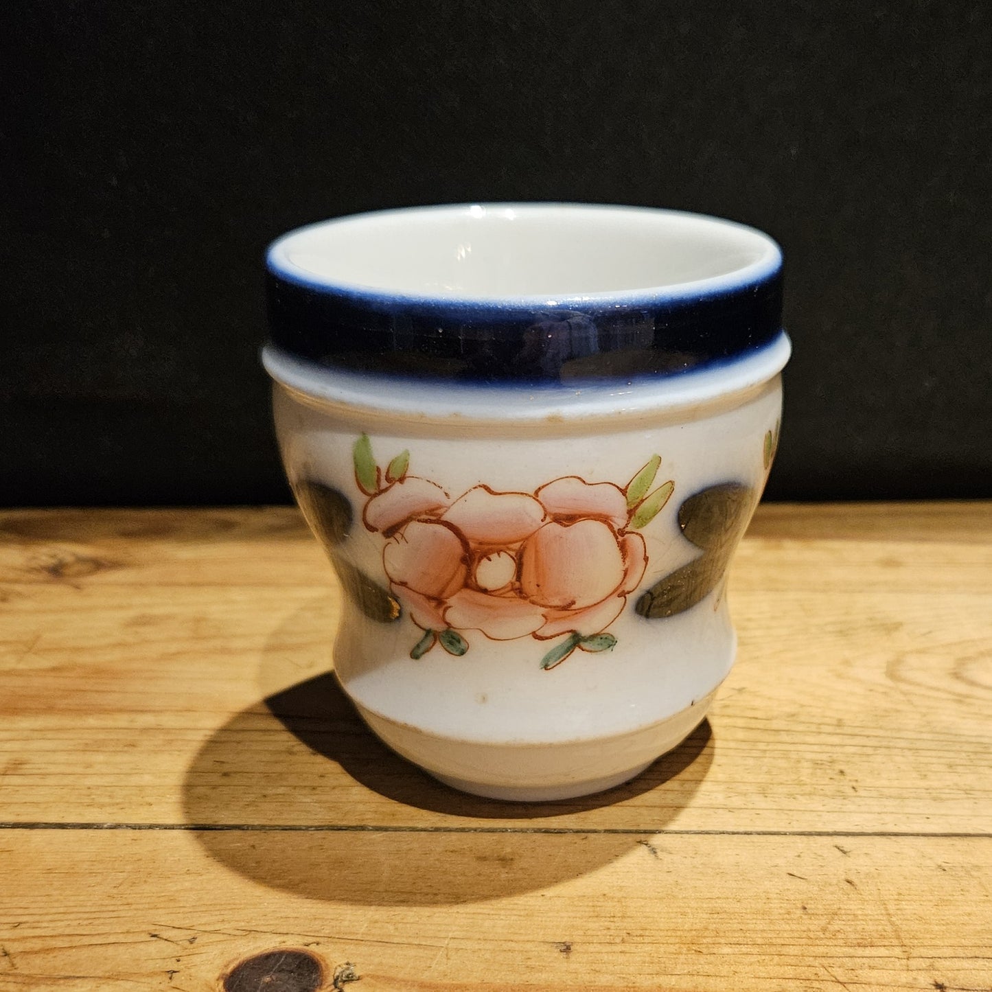 Vue arrière de la tasse à brûlot en faïence blanche, montrant le décor floral peint à la main avec une grande rose rose et des feuilles bleu-vert foncé, et le bandeau bleu cobalt sur le bord supérieur, fond noir.