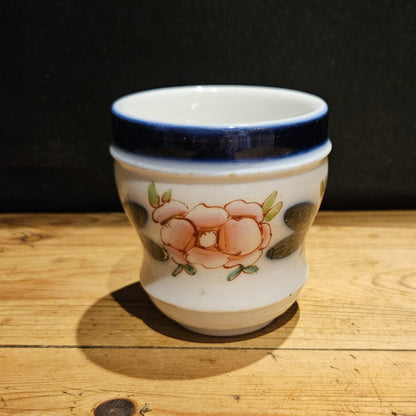 Vue arrière de la tasse à brûlot en faïence blanche, montrant le décor floral peint à la main avec une grande rose rose et des feuilles bleu-vert foncé, et le bandeau bleu cobalt sur le bord supérieur, fond noir.