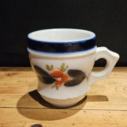 Vue de face de la tasse à brûlot en faïence blanche, anse à droite, avec bandeau bleu cobalt, décor de fleurs orangées et feuilles sombres peints à la main, posée sur une table en bois, fond noir.