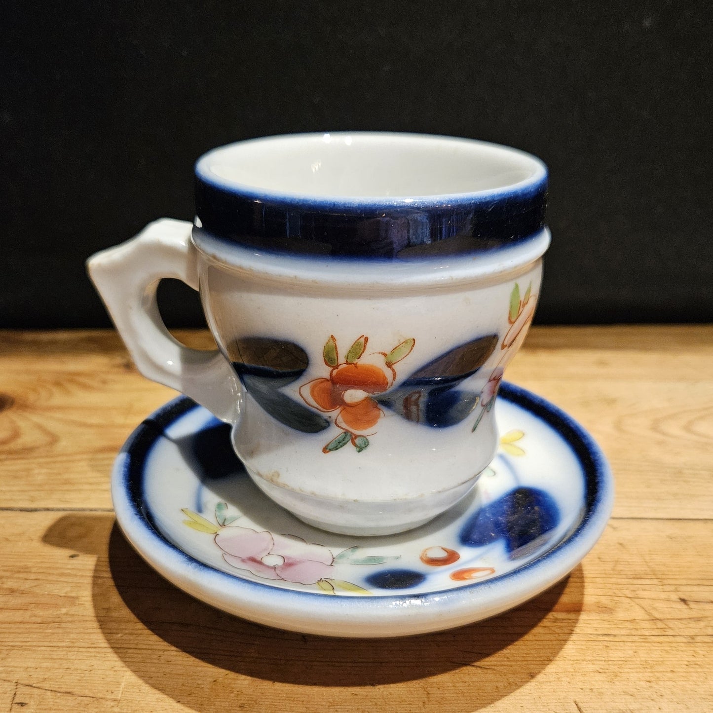 Ensemble tasse à brûlot et soucoupe en faïence blanche, avec bandeau bleu cobalt et décor floral peint à la main assorti, posés sur une table en bois brut, fond noir.