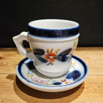 Ensemble tasse à brûlot et soucoupe en faïence blanche, avec bandeau bleu cobalt et décor floral peint à la main assorti, posés sur une table en bois brut, fond noir.