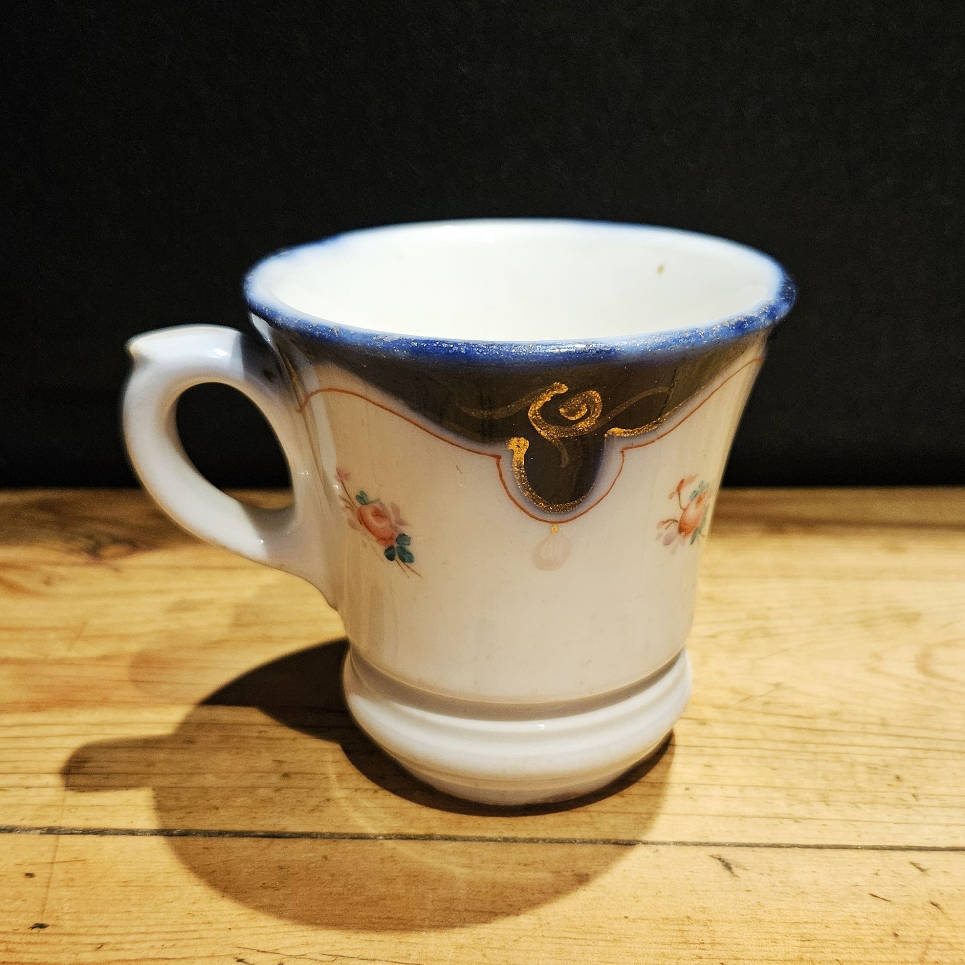 asse à brûlot ancienne en faïence blanche, avec large bandeau bleu cobalt sur le bord supérieur, volutes dorées, petites roses peintes à la main sur le corps blanc, anse en volute à gauche, posée sur une table en bois brut, fond noir.