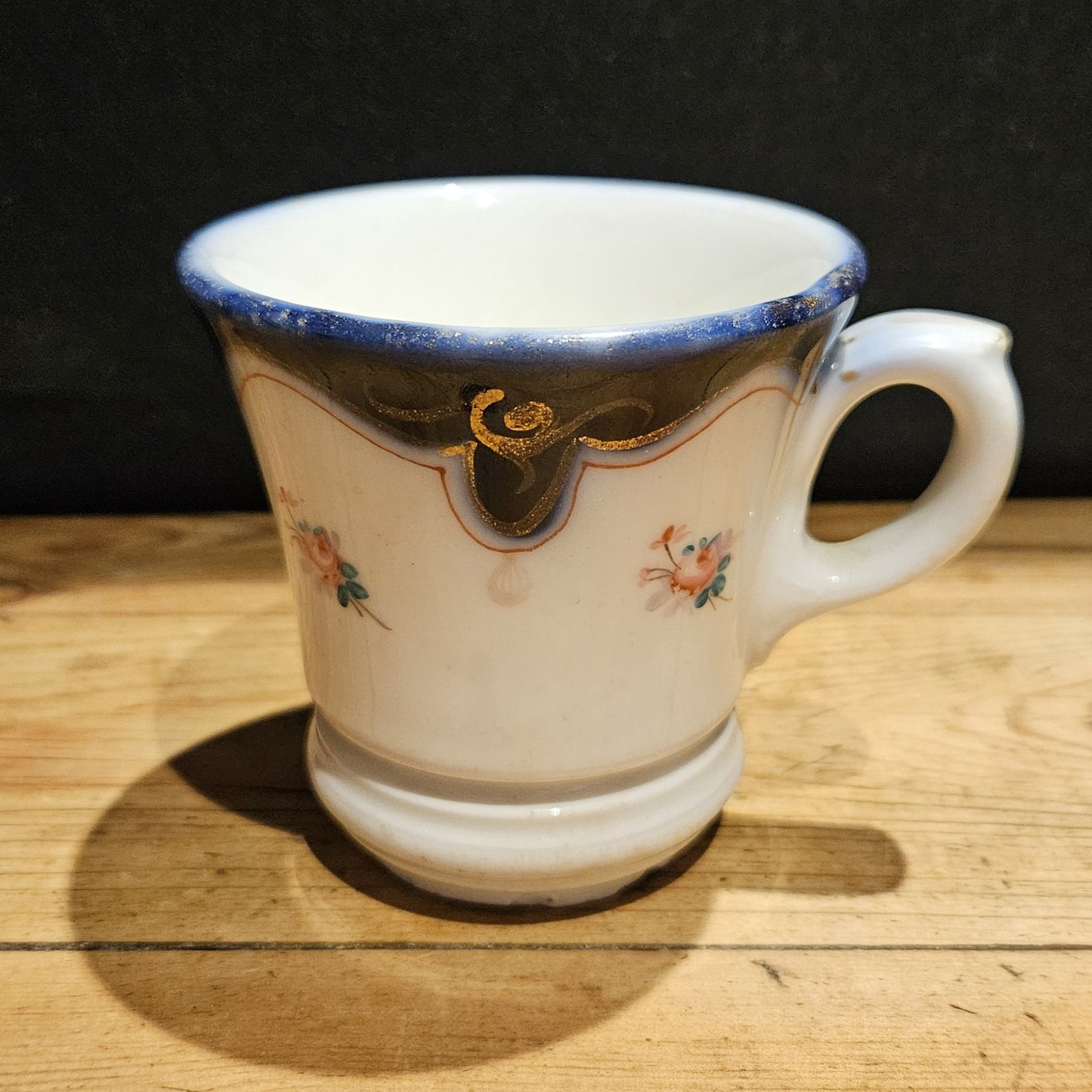 Vue arrière de la tasse à brûlot en faïence blanche, montrant l'anse en volute à droite, le bandeau bleu cobalt, les volutes dorées et les petites roses peintes à la main, posée sur une table en bois brut, fond noir.
