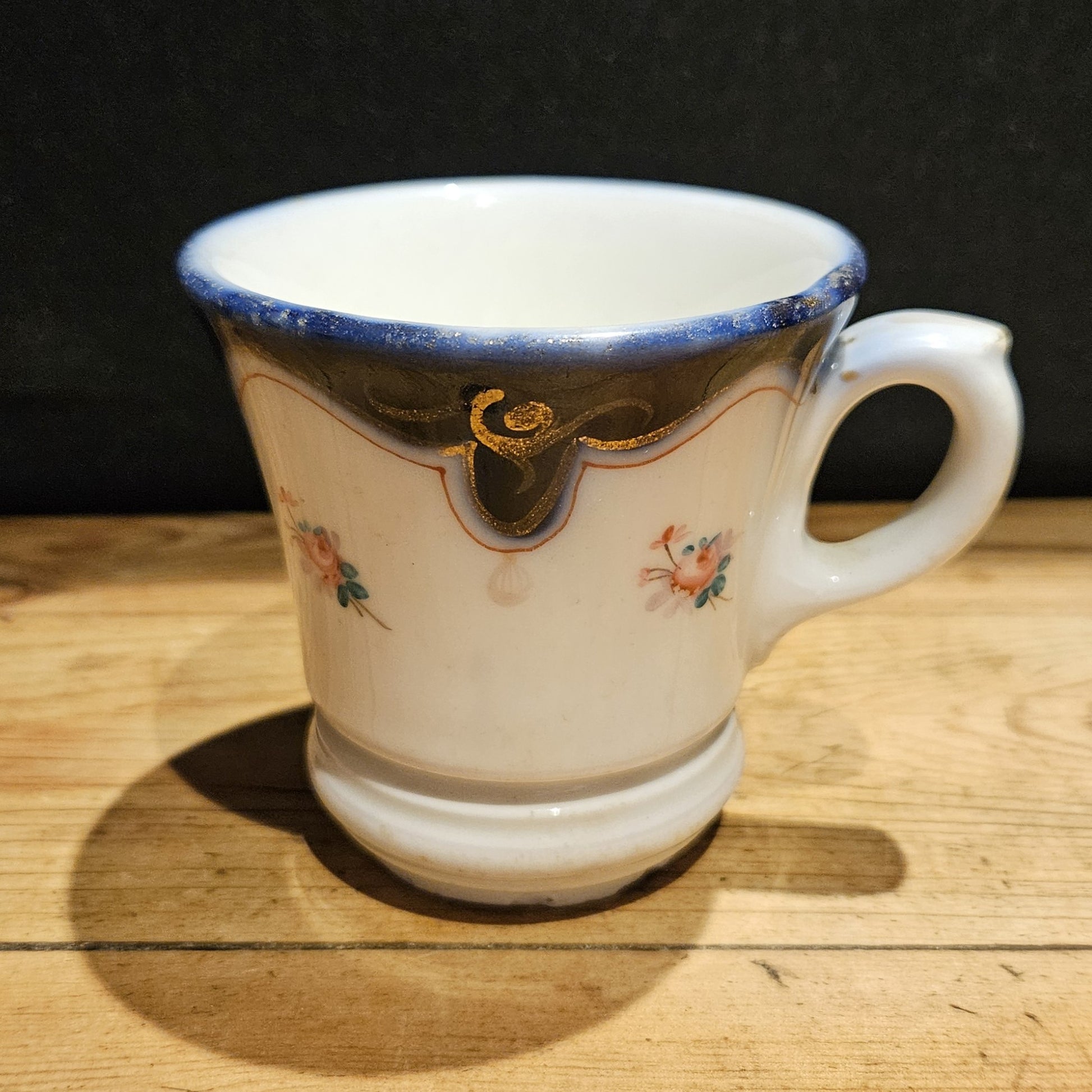 Vue arrière de la tasse à brûlot en faïence blanche, montrant l'anse en volute à droite, le bandeau bleu cobalt, les volutes dorées et les petites roses peintes à la main, posée sur une table en bois brut, fond noir.