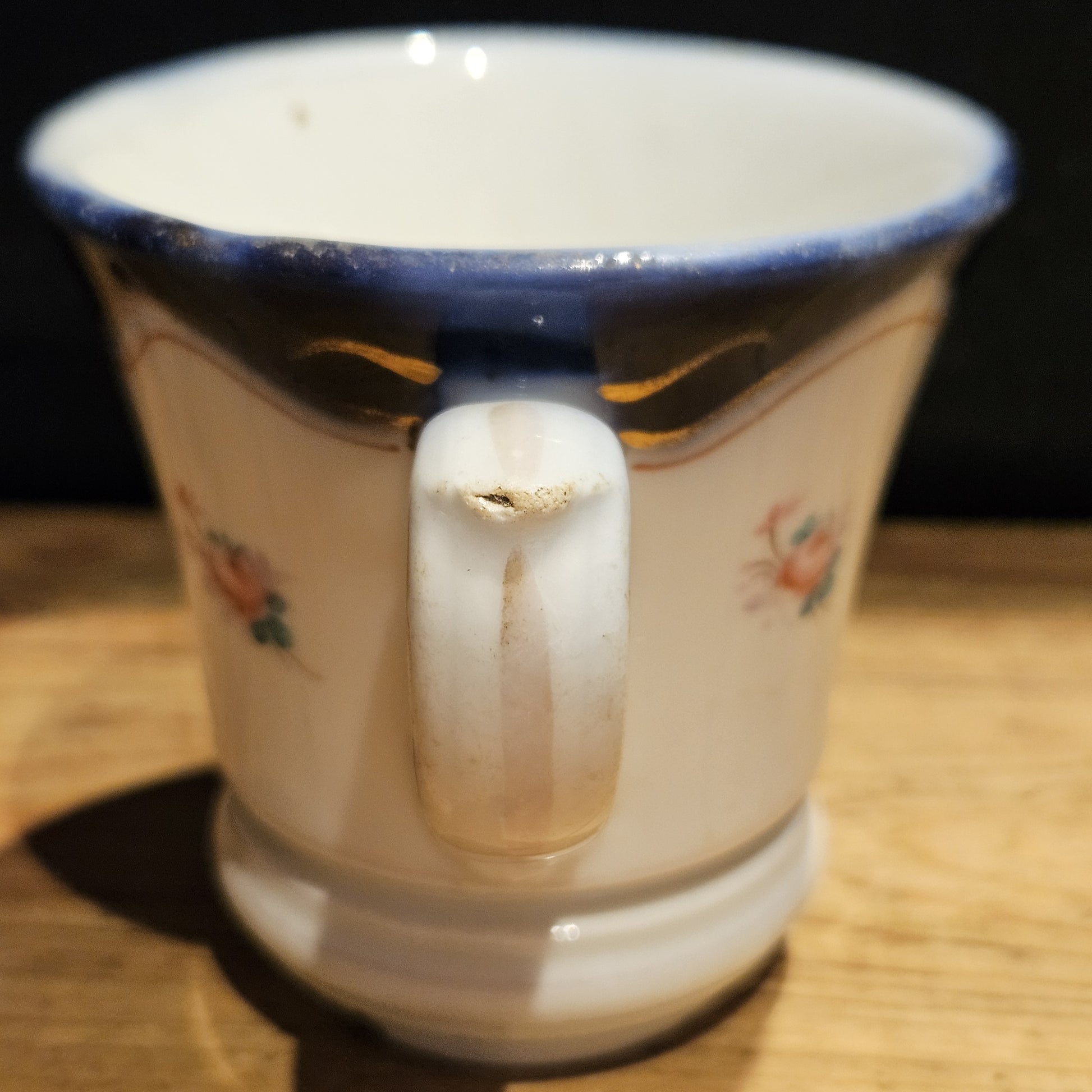 Gros plan sur la jonction supérieure de l'anse en volute de la tasse à brûlot en faïence blanche, montrant un petit trou d'usure visible, avec le bandeau bleu cobalt et les volutes dorées en arrière-plan.