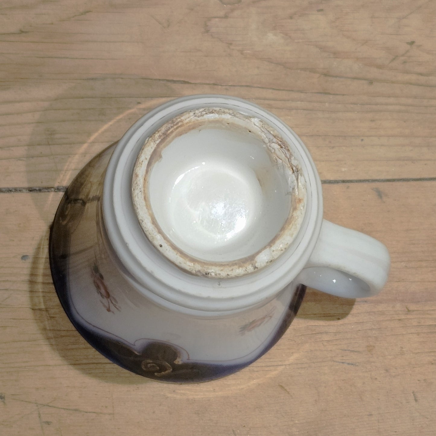 Vue du dessous de la tasse à brûlot en faïence blanche, montrant la base circulaire non émaillée avec patine d'ancienneté et légère usure, sans marque de potier visible, anse en volute visible sur le côté.