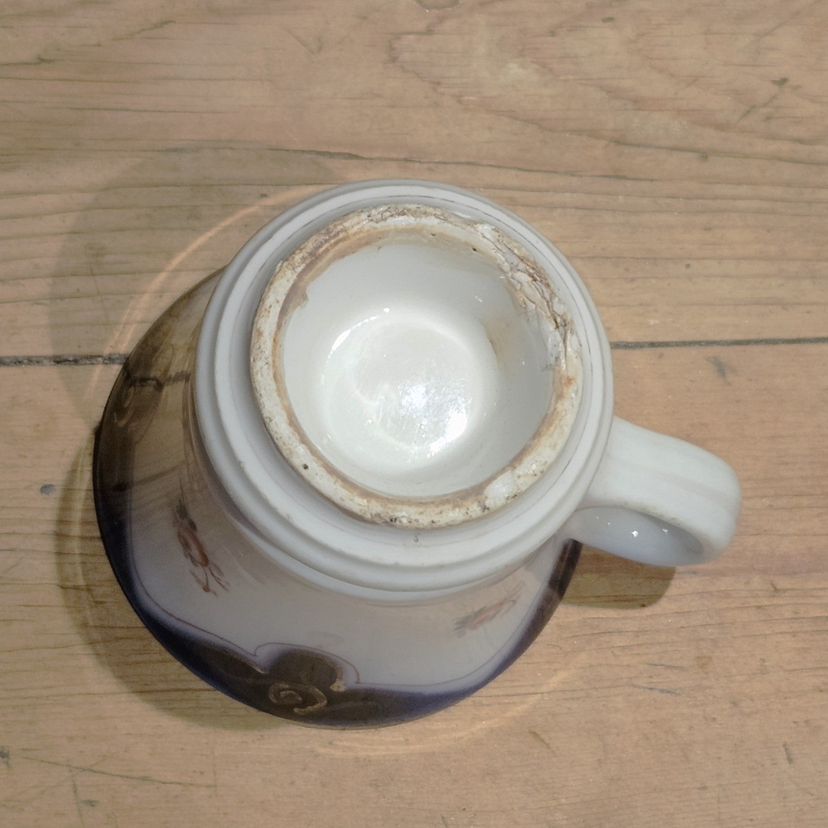 Vue du dessous de la tasse à brûlot en faïence blanche, montrant la base circulaire non émaillée avec patine d'ancienneté et légère usure, sans marque de potier visible, anse en volute visible sur le côté.