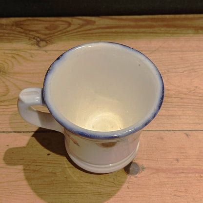 Vue plongeante de l'intérieur de la tasse à brûlot en faïence blanche, montrant l'émail blanc légèrement crème, le bord avec bandeau bleu cobalt usé, et l'anse en volute visible à gauche, posée sur une table en bois.