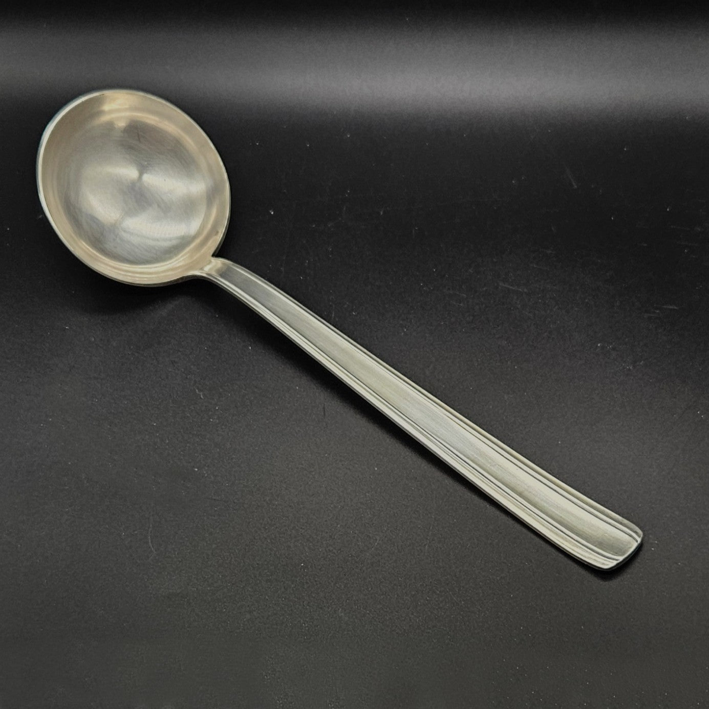 Louche à potage en métal argenté Moderniste, 30,7 cm, vue de face sur fond noir, montrant la cuillère ronde et profonde avec jonction manche/cuillère fluide et le manche plat rectiligne à double filet latéral fin.