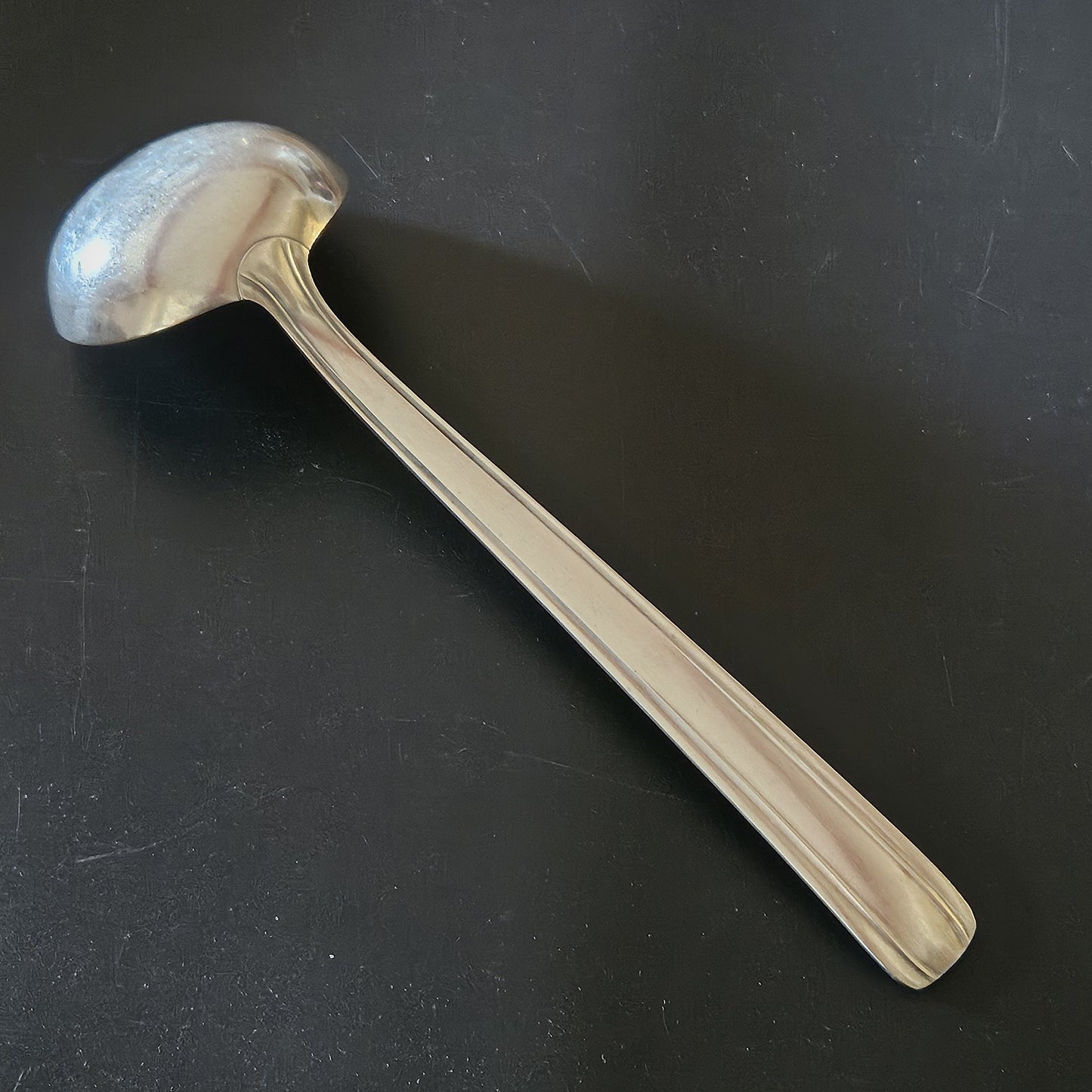 Louche à potage en métal argenté Moderniste, 30,7 cm, vue de dos sur fond noir, montrant le dos de la cuillère ronde et le manche plat rectiligne à double filet latéral fin avec extrémité carrée légèrement arrondie.
