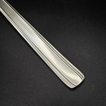 Gros plan sur l'extrémité carrée légèrement arrondie du manche de la louche à potage en métal argenté Moderniste, montrant le double filet latéral fin courant sur toute la longueur du manche plat et rectiligne, sur fond noir.