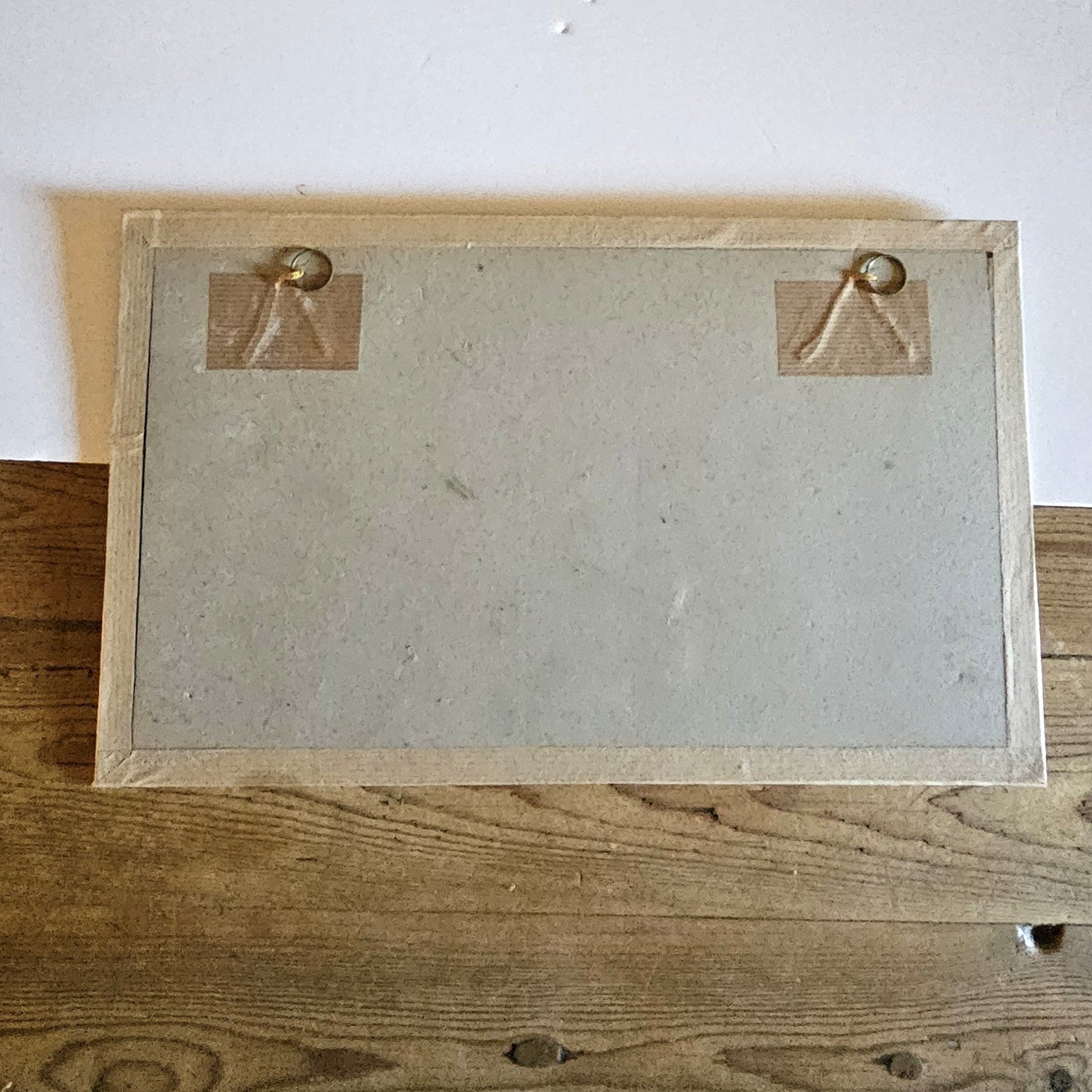 Vue du dos du cadre de l'aquarelle de Paul Friesé, montrant un fond en carton gris avec deux anneaux de suspension dorés fixés par du scotch kraft, dans un cadre en papier texturé beige/doré, posé sur une table en bois brut.