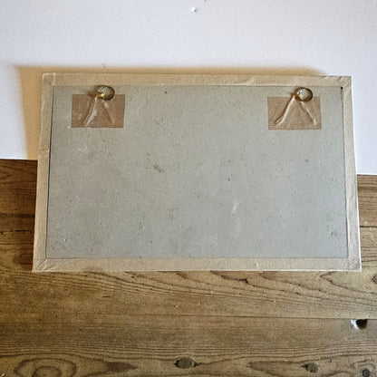 Vue du dos du cadre de l'aquarelle de Paul Friesé, montrant un fond en carton gris avec deux anneaux de suspension dorés fixés par du scotch kraft, dans un cadre en papier texturé beige/doré, posé sur une table en bois brut.