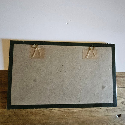 Vue du dos du cadre de l'aquarelle de Paul Friesé, montrant un fond en carton gris avec deux anneaux de suspension dorés fixés par du scotch kraft sur des étiquettes en carton, dans un cadre en papier texturé noir, posé sur une table en bois brut.