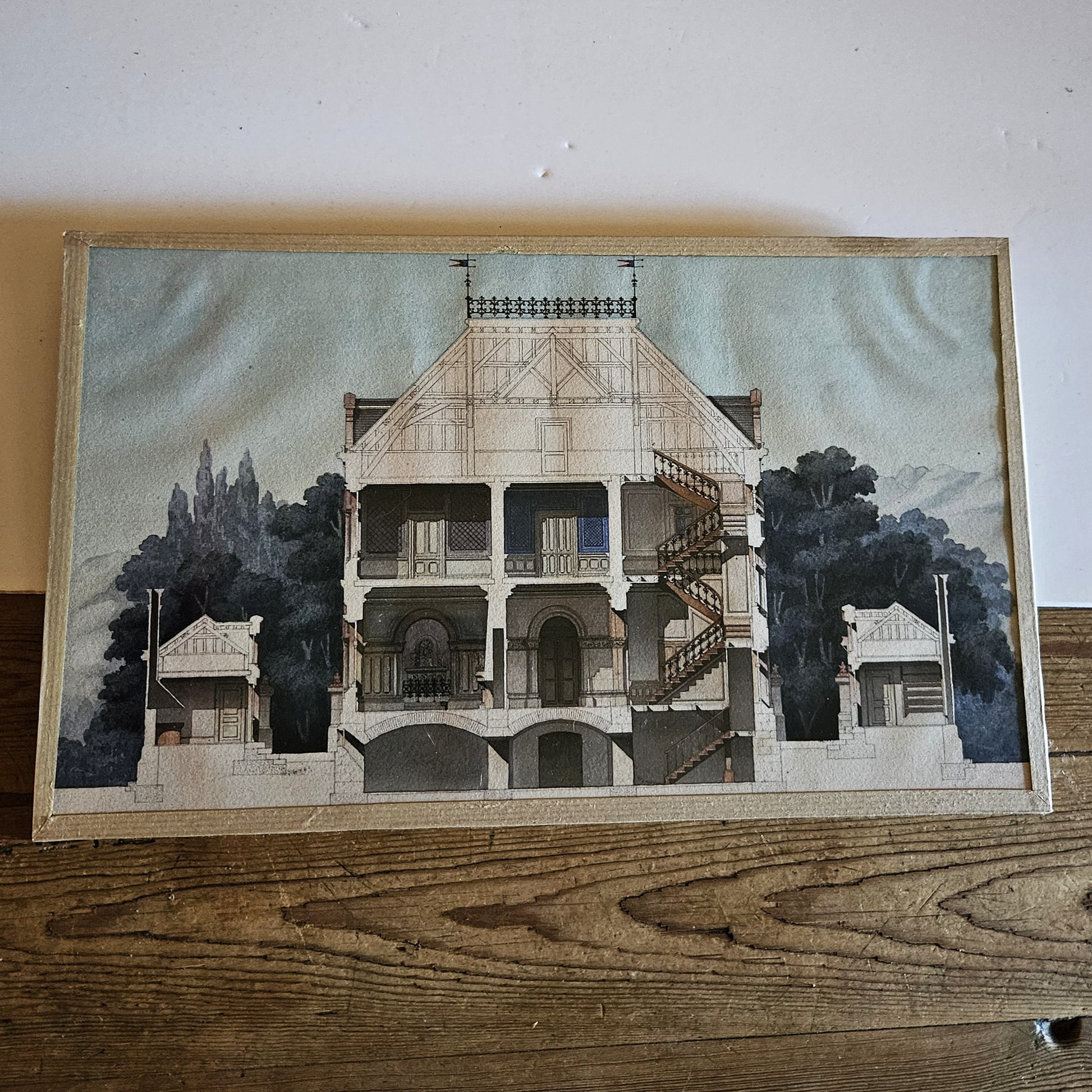 Aquarelle originale encadrée attribuée à Paul Friesé, représentant la coupe transversale d'une villa bourgeoise éclectique avec charpente apparente en bois à fermes triangulées, balustrade en fer forgé à fleurs de lys et girouettes en fanion au faîtage, double escalier hélicoïdal monumental, arcades en plein cintre en brique et pavillon de jardin à droite, sur fond de végétation dense au lavis gris-bleu, dans un cadre en papier texturé beige/crème, posée contre un mur beige.