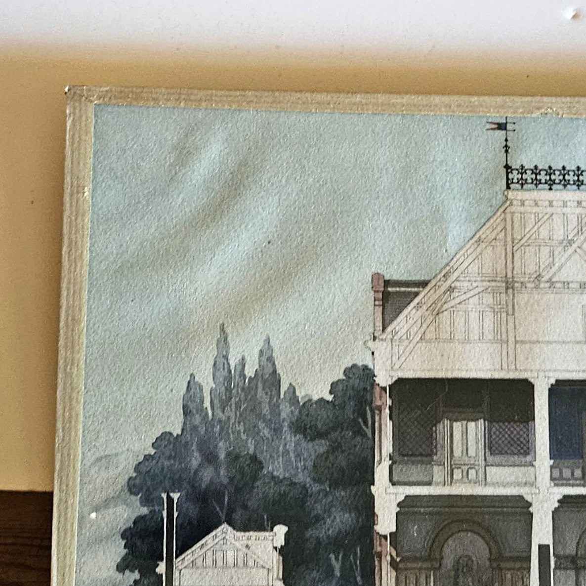 Détail de l'aquarelle de Paul Friesé, montrant le coin supérieur gauche de la composition avec la charpente apparente en bois à fermes triangulées de la villa bourgeoise, la balustrade en fer forgé à fleurs de lys et girouettes en fanion au faîtage, et la végétation au lavis gris-bleu, dans un cadre en papier texturé beige/crème.