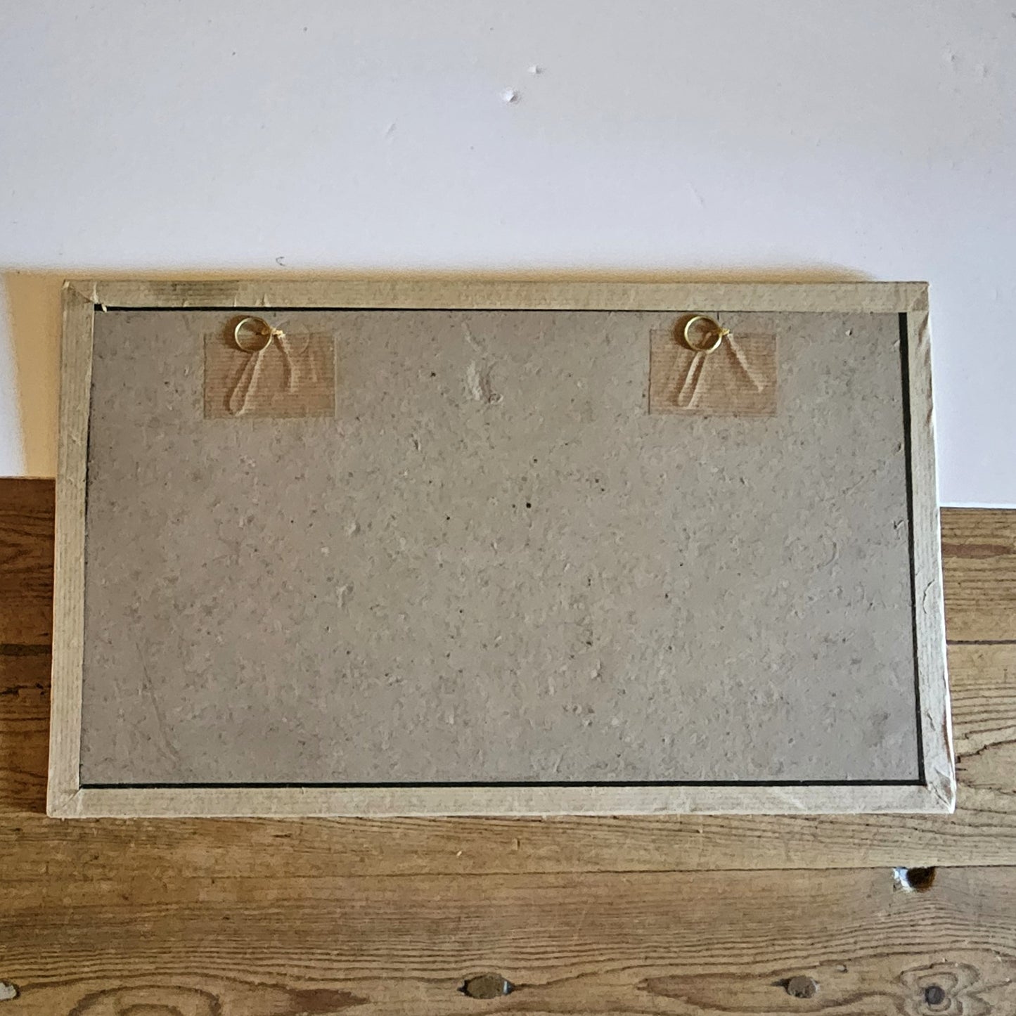 Vue du dos du cadre de l'aquarelle de Paul Friesé, montrant un fond en carton gris avec deux anneaux de suspension dorés fixés par du scotch kraft sur des étiquettes en carton ondulé, dans un cadre en papier texturé beige/crème, posé sur une table en bois brut.