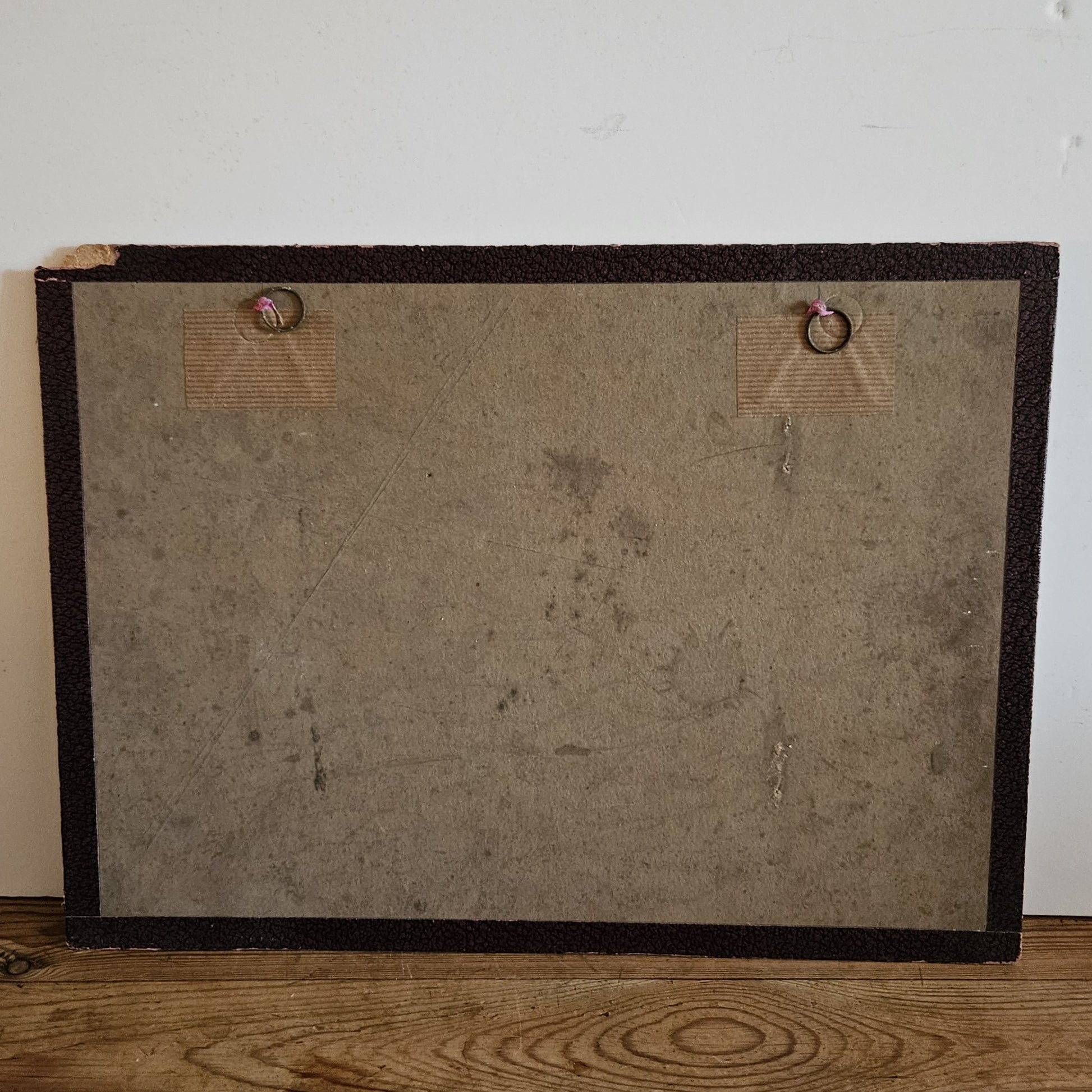 Vue du dos du cadre de l'aquarelle de Paul Friesé, montrant un fond en carton brun avec deux anneaux de suspension fixés par des étiquettes en carton, posé sur un parquet en bois brut.