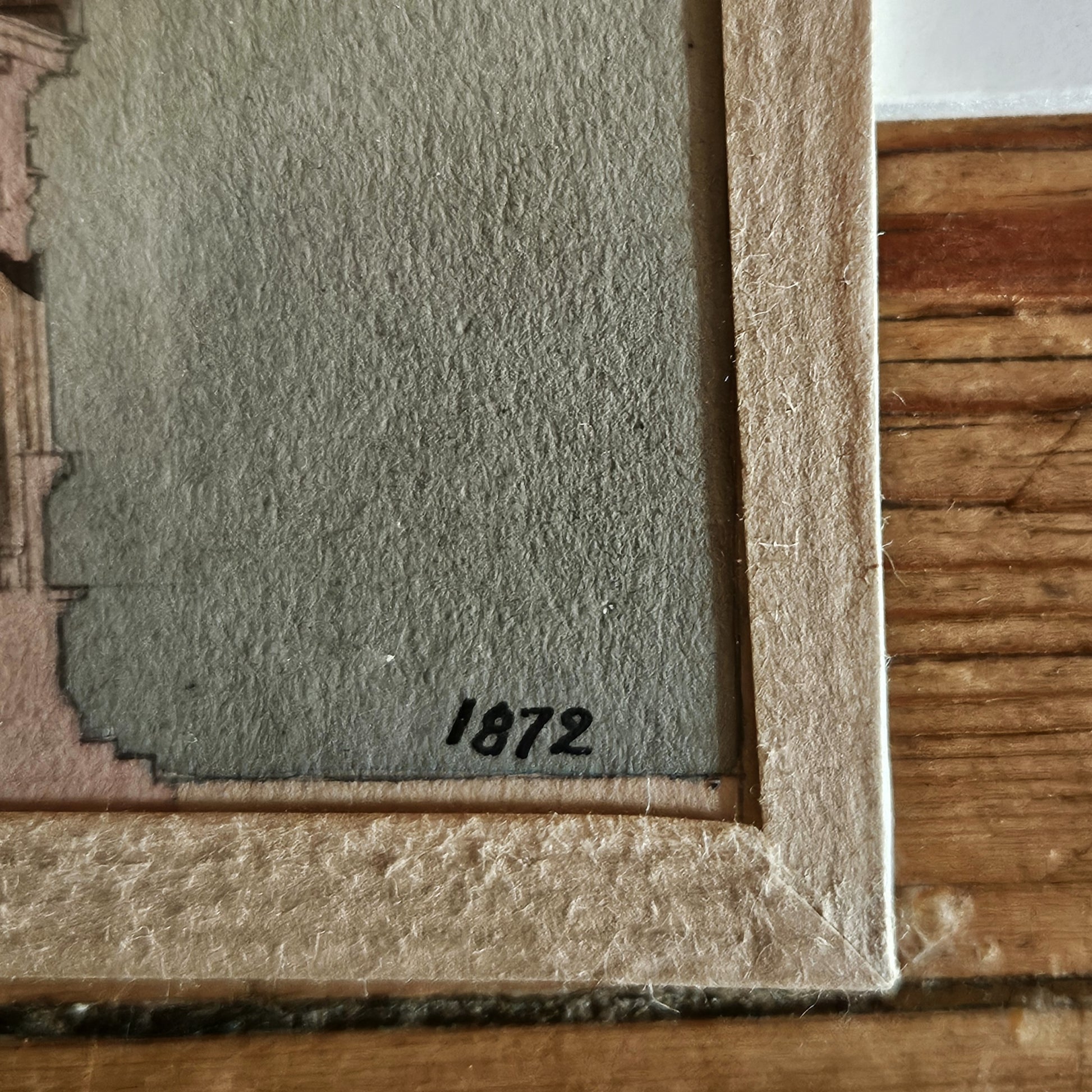 Gros plan sur le coin inférieur droit de l'aquarelle de Paul Friesé, montrant la date « 1872 » inscrite à l'encre noire sur le papier vergé beige, avec le cadre en papier texturé beige et le bord de la table en bois brut visibles.