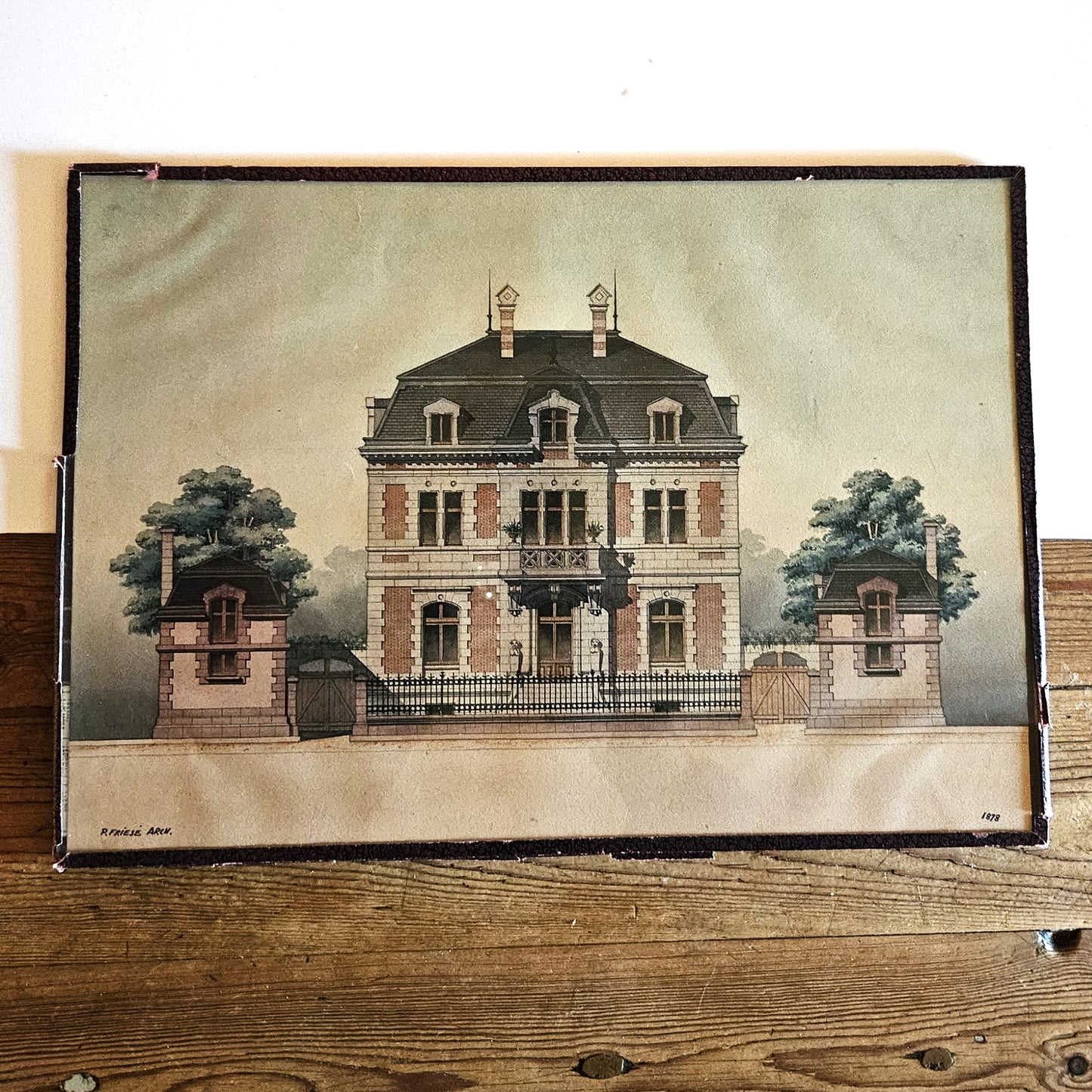 Aquarelle originale encadrée de Paul Friesé, représentant la façade d'une villa bourgeoise Napoléon III en brique rouge et pierre de taille avec toit à la Mansart, balcon central et deux pavillons d'entrée, signée P. FRIESE ARCH. et datée 1878, dans un cadre en papier texturé brun foncé, posée sur un parquet en bois brut.