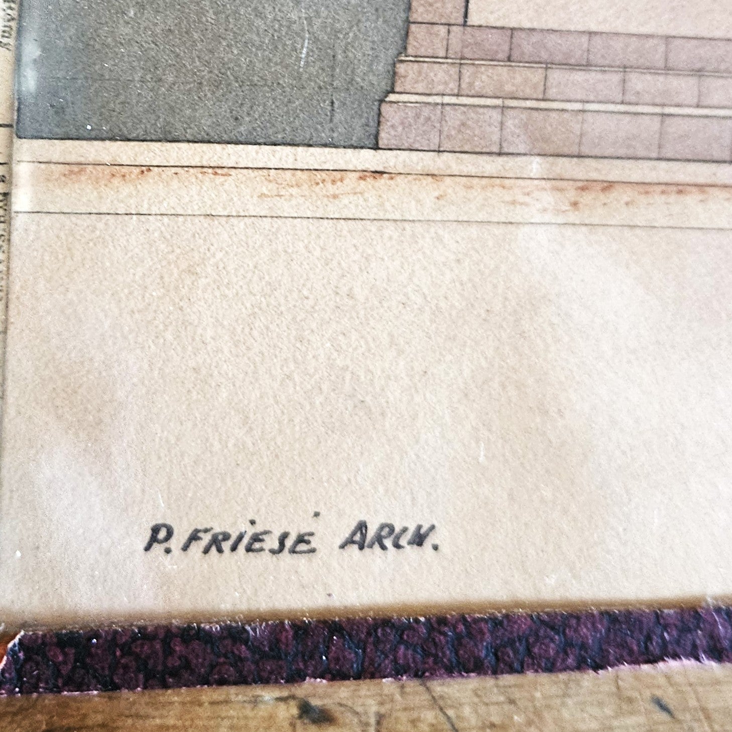 Gros plan sur la signature manuscrite « P. FRIESE ARCH. » inscrite en bas à gauche de l'aquarelle, sur le bord blanc du papier vergé, dans un cadre en papier texturé brun foncé, posée sur un parquet en bois brut.
