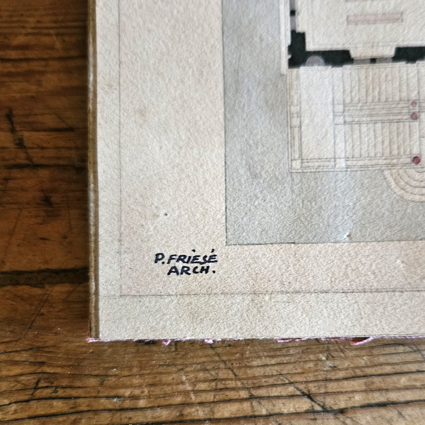 Gros plan sur le coin inférieur gauche du dessin d'architecture de Paul Friesé, montrant la signature manuscrite « P. FRIESÉ ARCH. » à l'encre noire sur le papier vergé beige, avec le bord du carton d'époque visible, posé sur une table en bois brut.