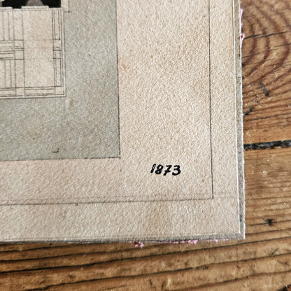 Gros plan sur le coin inférieur droit du dessin d'architecture de Paul Friesé, montrant la date « 1873 » inscrite à l'encre noire sur le papier vergé beige, avec le bord du carton d'époque visible, posé sur une table en bois brut.