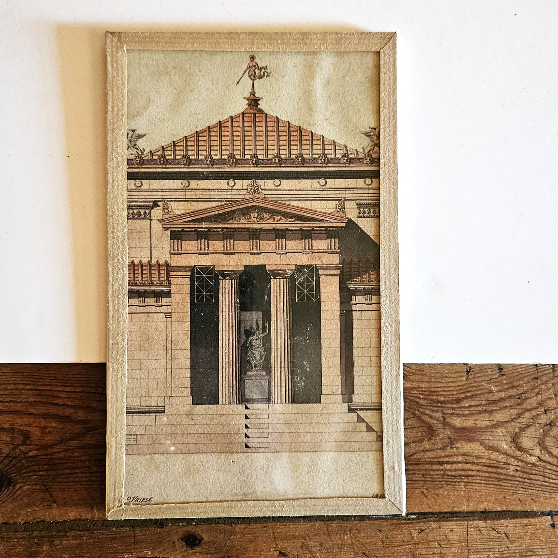 Aquarelle originale polychrome encadrée de Paul Friesé, représentant la façade d'un édifice public néo-grec monumental avec grand fronton, girouette dorée en forme de Victoire, frise polychrome et portique à colonnes corinthiennes, dans un cadre en papier texturé beige, posée sur une table en bois brut.