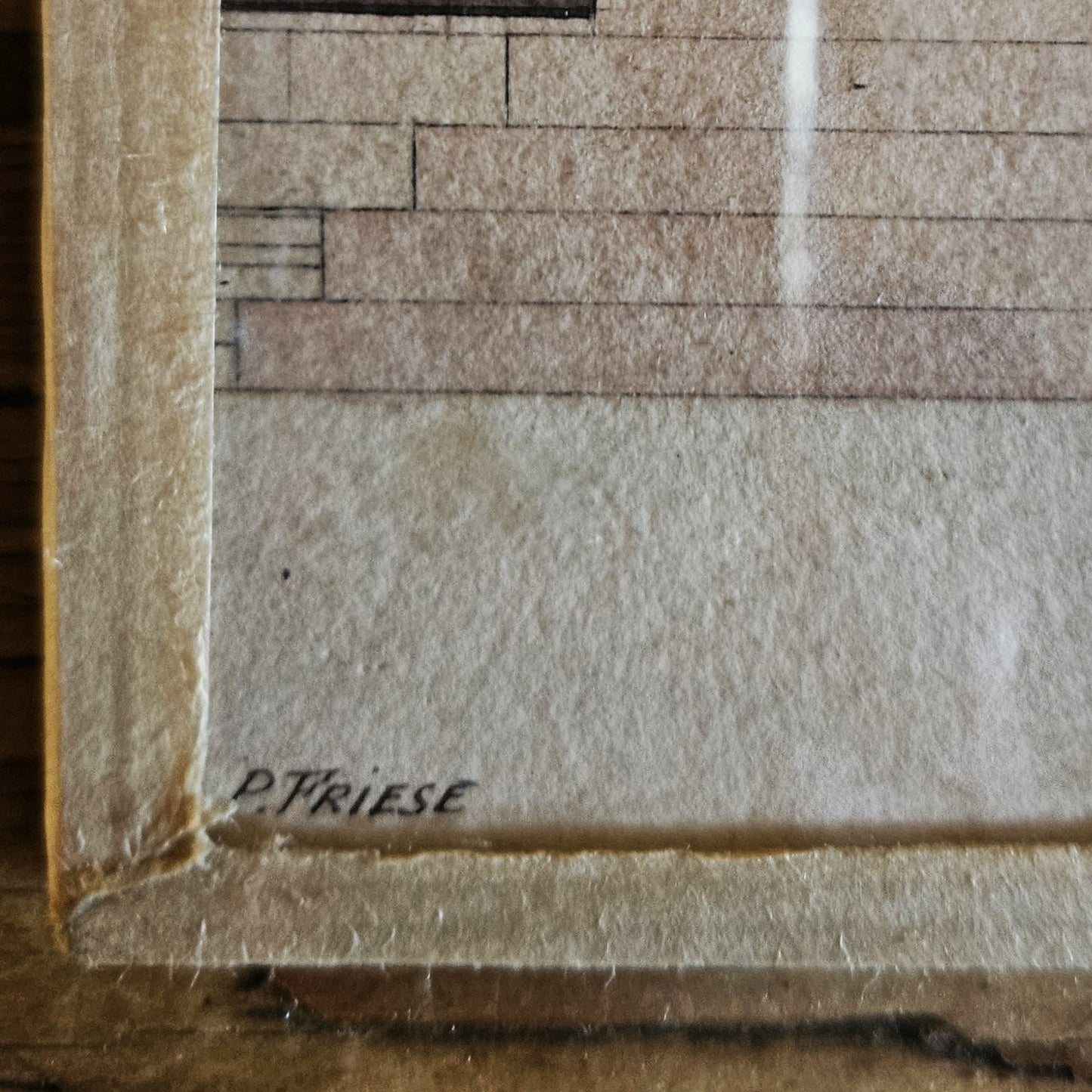 Gros plan sur le coin inférieur gauche de l'aquarelle de Paul Friesé, montrant la signature manuscrite « P. FRIESE » à l'encre noire sur le papier vergé beige, avec le bord du cadre en papier texturé beige visible, posé sur une table en bois brut.