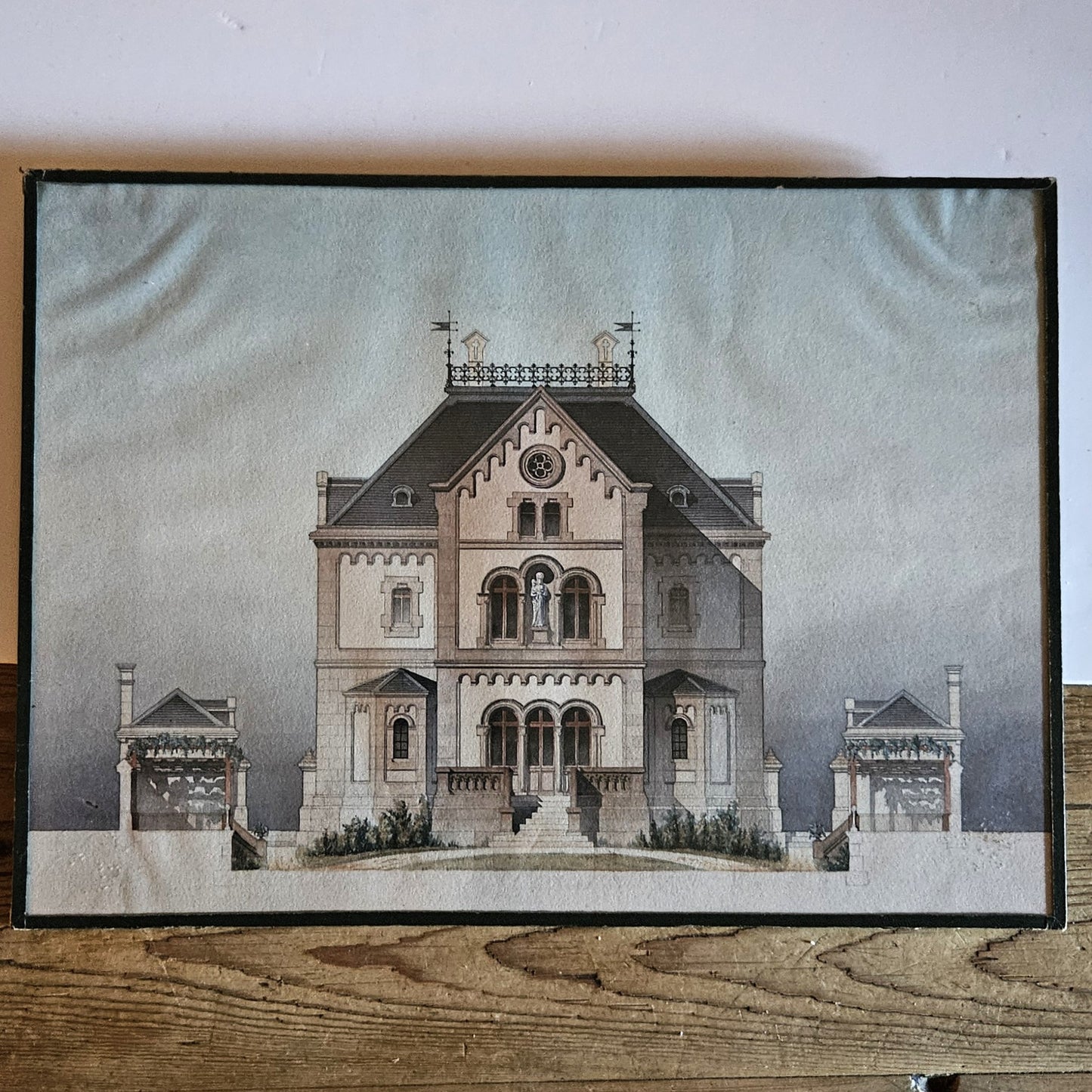 Vue d'ensemble de l'aquarelle originale encadrée attribuée à Paul Friesé, représentant la façade d'un édifice religieux néo-roman avec statue de la Vierge à l'Enfant, perron monumental et pavillons latéraux, dans un sous verre entouré d'un papier noir d'origine, posée sur une table en bois brut.