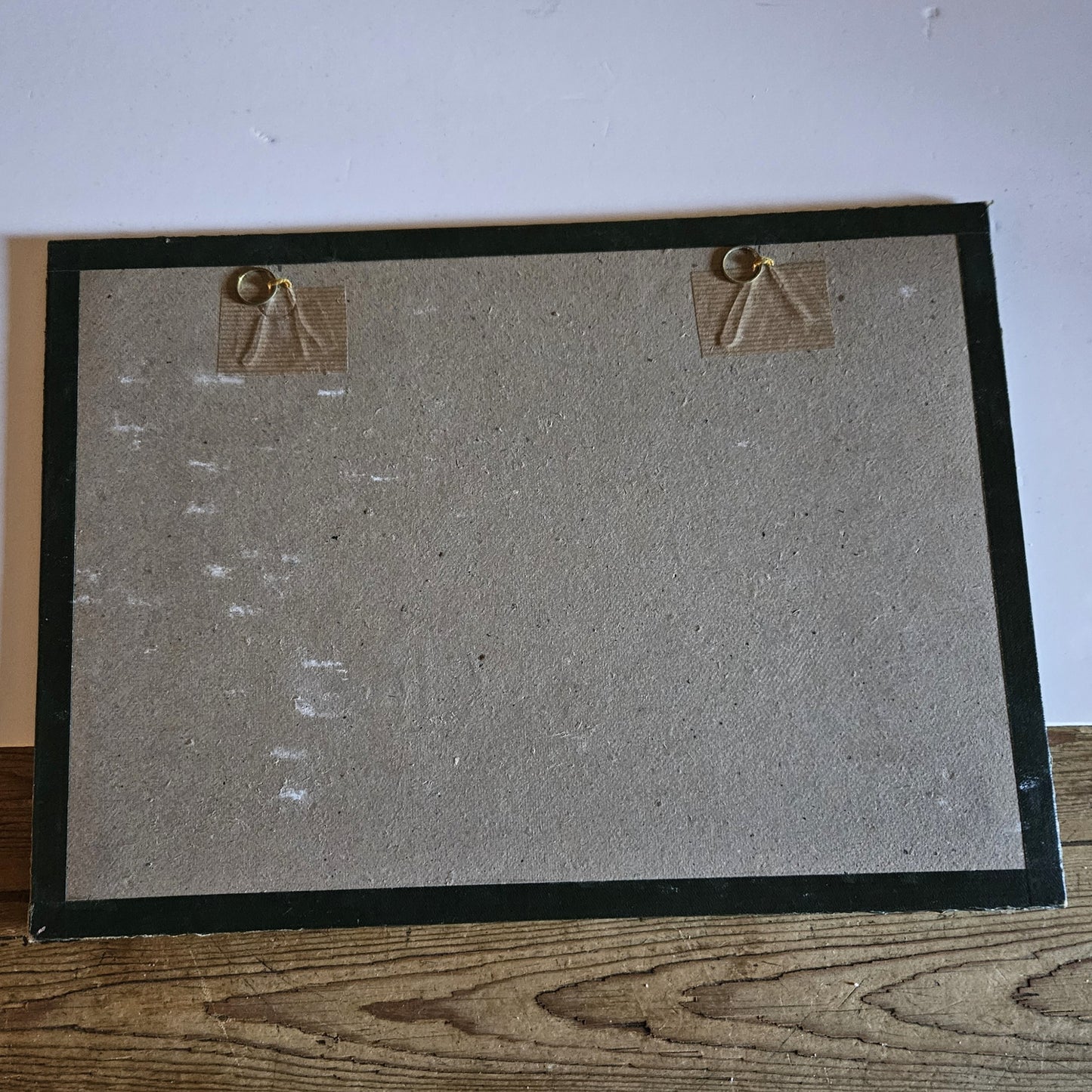 Vue du dos du cadre de l'aquarelle attribuée à Paul Friesé, montrant un fond en carton gris avec deux anneaux de suspension dorés fixés par des étiquettes en carton ondulé, dans un sou-verre entouré d'un papier noir d'origine, posé sur une table en bois brut.