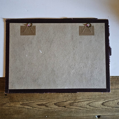 Vue du dos du cadre de l'aquarelle de Paul Friesé, montrant un fond en carton gris avec deux anneaux de suspension fixés par des étiquettes en carton kraft posé sur une table en bois brut.