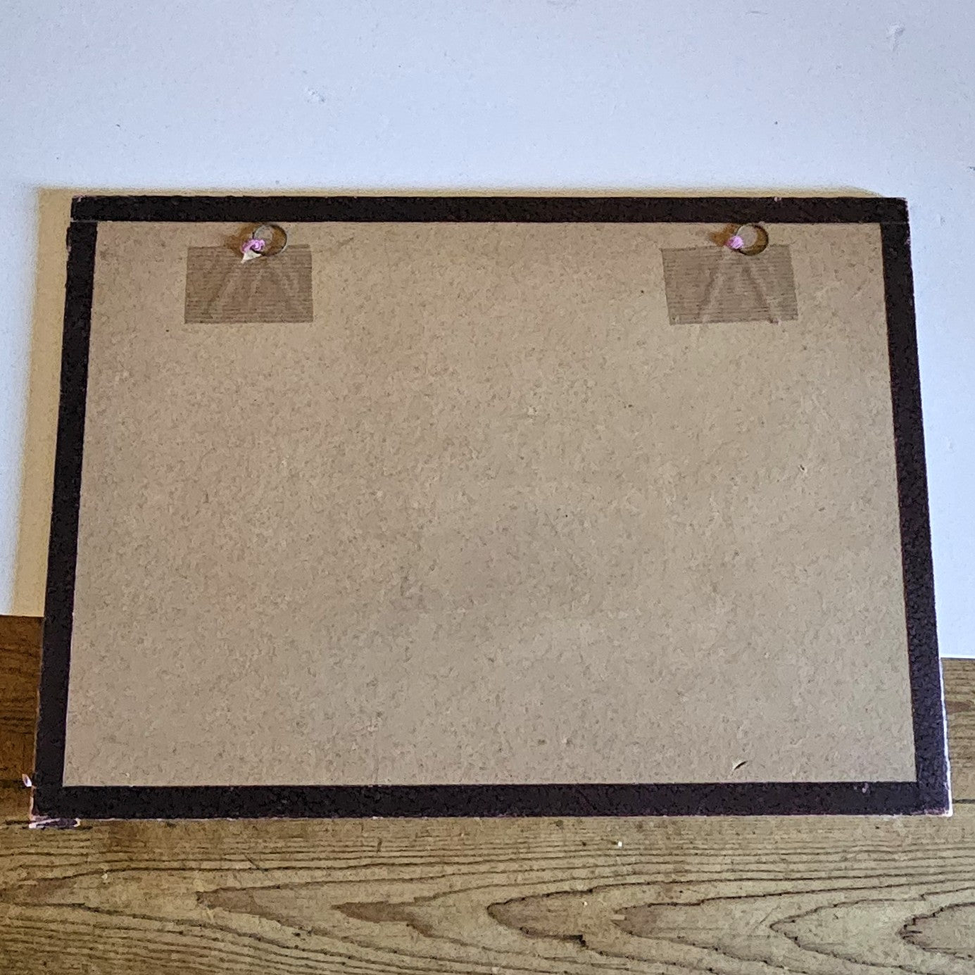 Vue du dos du cadre de l'aquarelle attribuée à Paul Friesé, montrant un fond en carton beige avec deux anneaux de suspension fixés par du scotch kraft, dans un cadre en papier texturé noir, posé sur un parquet en bois brut.