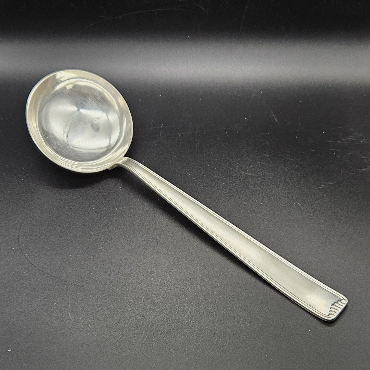 Louche à potage en métal argenté Art Déco, 31 cm, vue de face sur fond noir, montrant la cuillère ronde et profonde avec bourrelet de jonction et le manche rectangulaire à double filet latéral avec extrémité en éventail à cannelures.