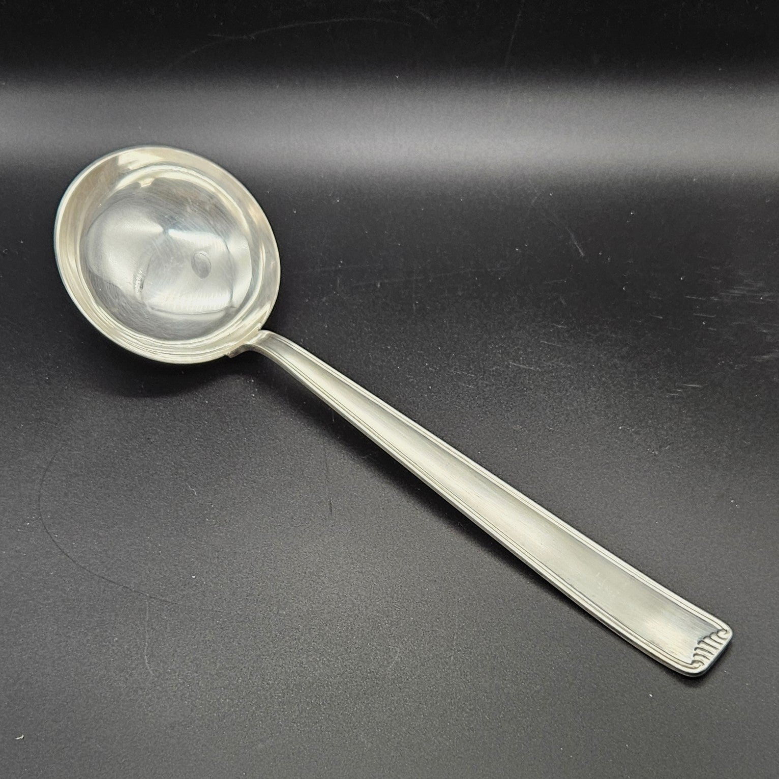 Louche à potage en métal argenté Art Déco, 31 cm, vue de face sur fond noir, montrant la cuillère ronde et profonde avec bourrelet de jonction et le manche rectangulaire à double filet latéral avec extrémité en éventail à cannelures.