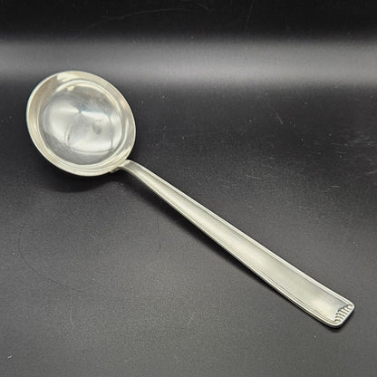 Louche à potage en métal argenté Art Déco, 31 cm, vue de face sur fond noir, montrant la cuillère ronde et profonde avec bourrelet de jonction et le manche rectangulaire à double filet latéral avec extrémité en éventail à cannelures.