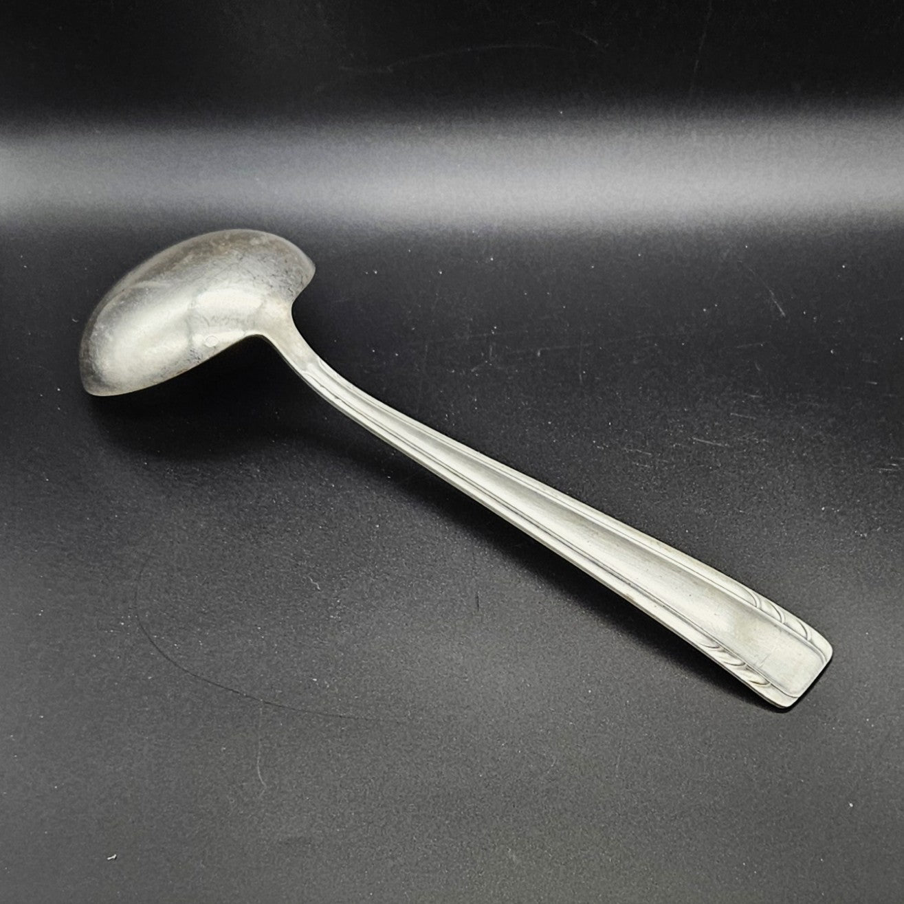 Louche à potage en métal argenté Art Déco, 30 cm, vue de dos sur fond noir, montrant le dos de la cuillère ronde et le manche rectangulaire effilé à double filet latéral avec extrémité arrondie à fines cannelures en arc de cercle.
